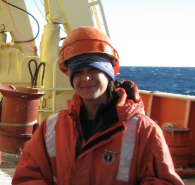 Michelle poses outside on a research vessel ship, wearing a hard hat and a big orange jacket