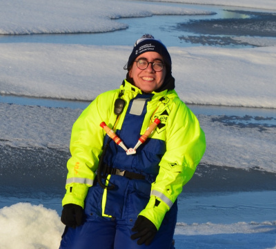 Alex (a woman with dark brown hair and tanned skin, wearing a bobble hat, bright yellow jacket and blue overalls) kneels on seal ice doing science. 