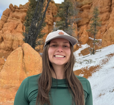 Mikaela Williams stands in front of bright orange rock with snow on it, wearing a green long-sleeved top and a white hat, grinning at the camera