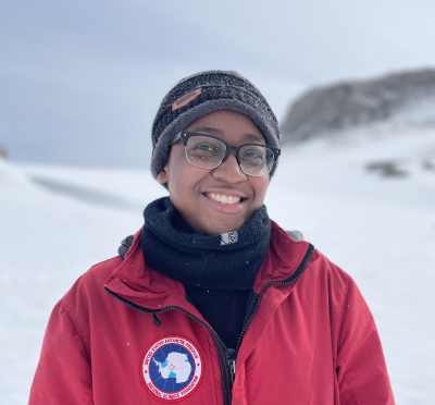 Maya Thomas stands in Antarctica wearing a warm hat, glasses, a buff and a red 'USAP NSF' jacket