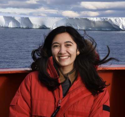 Asmara Lehrmann stands on a boat in front of a big ice shelf in Antarctica with ocean in between. Asmara wears a big red jacket and smiles at the camera. 