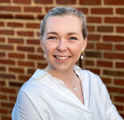 Marion McKenzie (a woman with dark blonde hair and light skin wearing earrings and a white blouse) smiles at the camera in front of a red brick wall