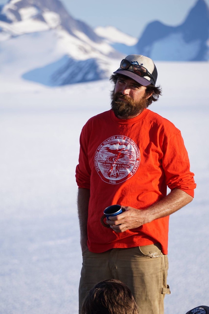 Chris McNeil (man with fair skin and dark brown hair and beard) wearing a hat and a red shirt with the Juneau Icefield Research Program logo on it stands in front of a glaciated mountain scene holding a mug of coffee.
