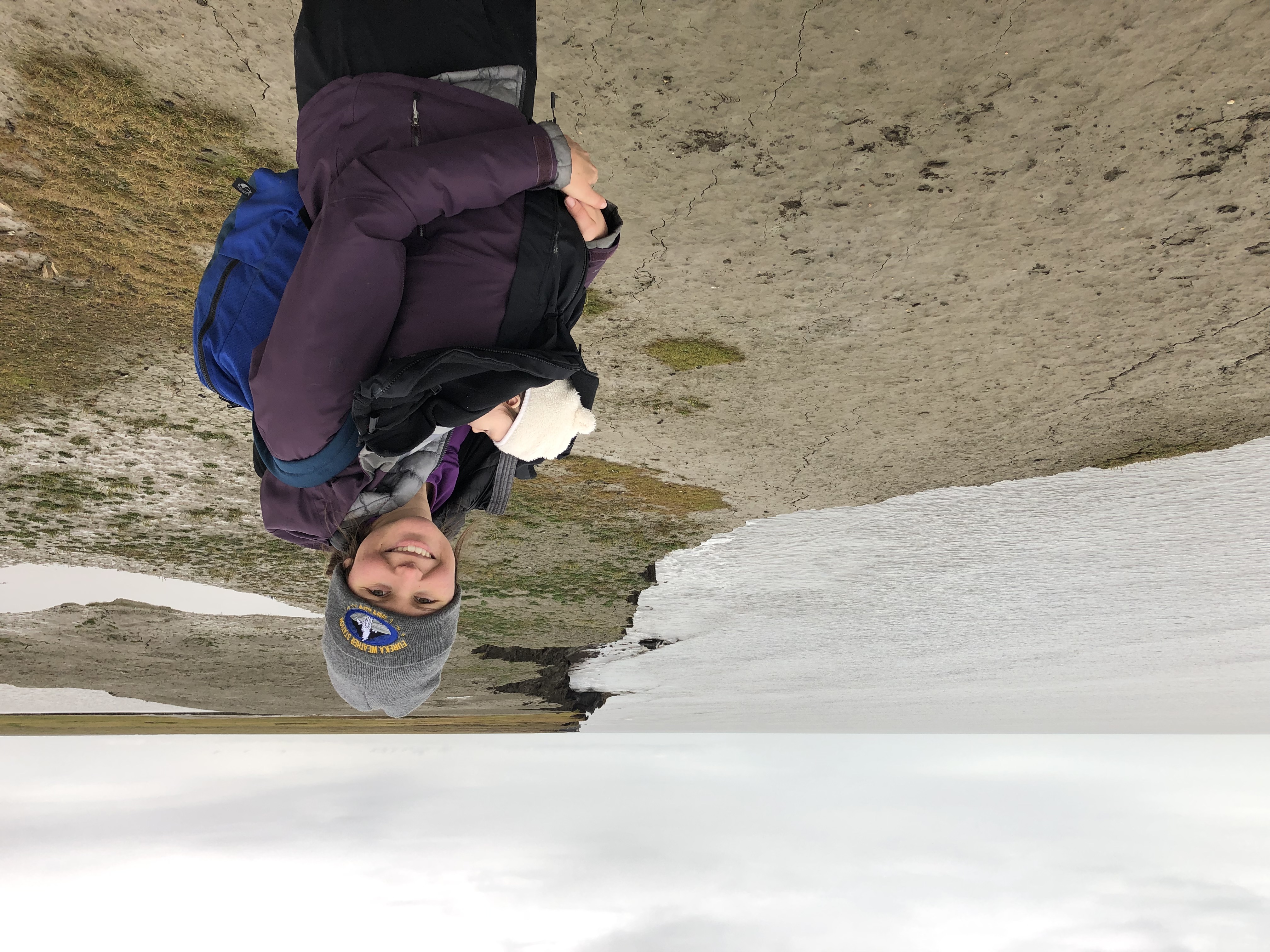 Melissa smiles at the camera at Drew Point, Alaska, with her baby swaddled at her front.