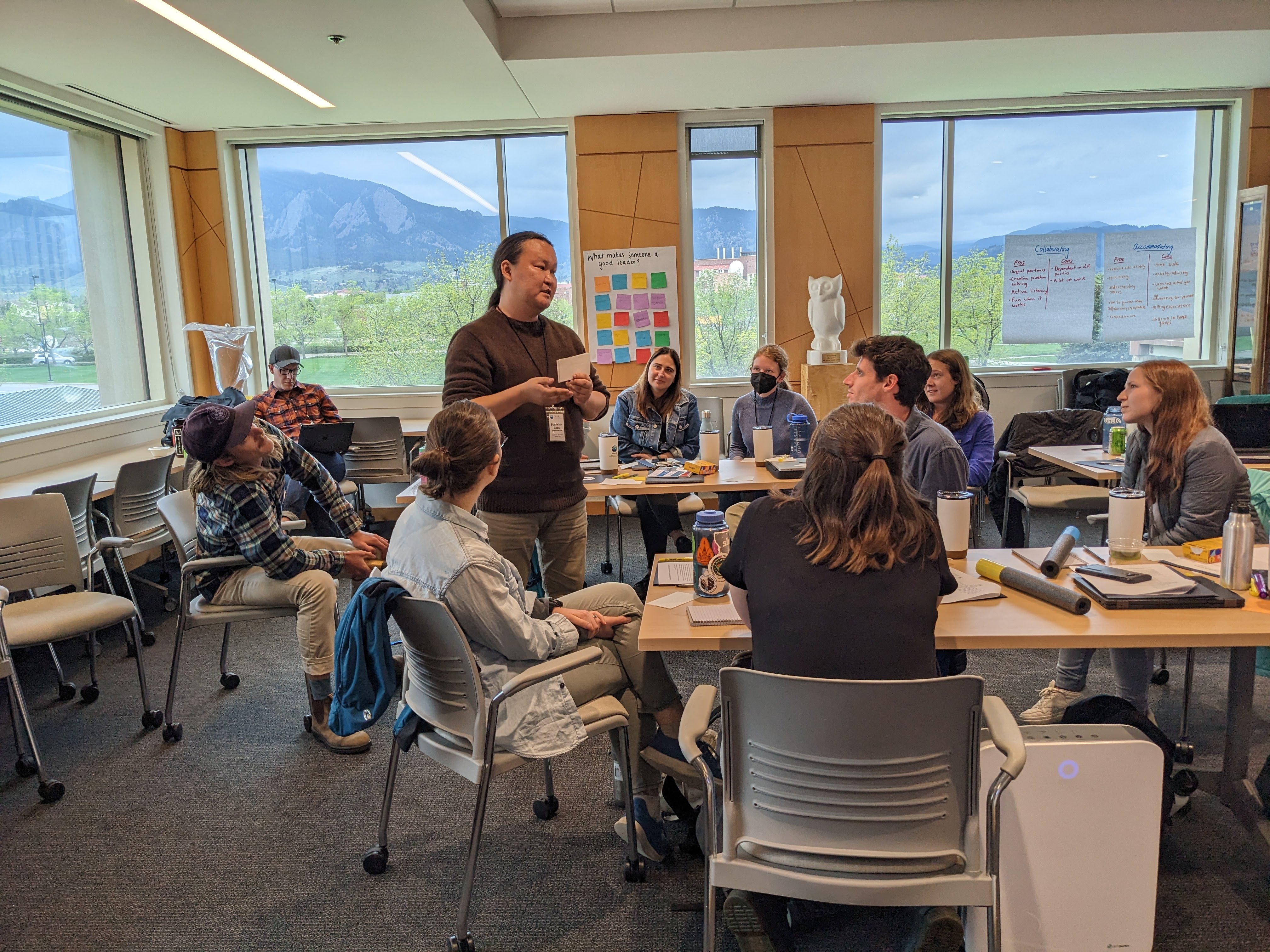 Postdoctoral researcher practice describing their research with using only the most common words in the English language. A person stands and speaks to nine other seated people inside a conference room. The Boulder flat irons are in the background. 