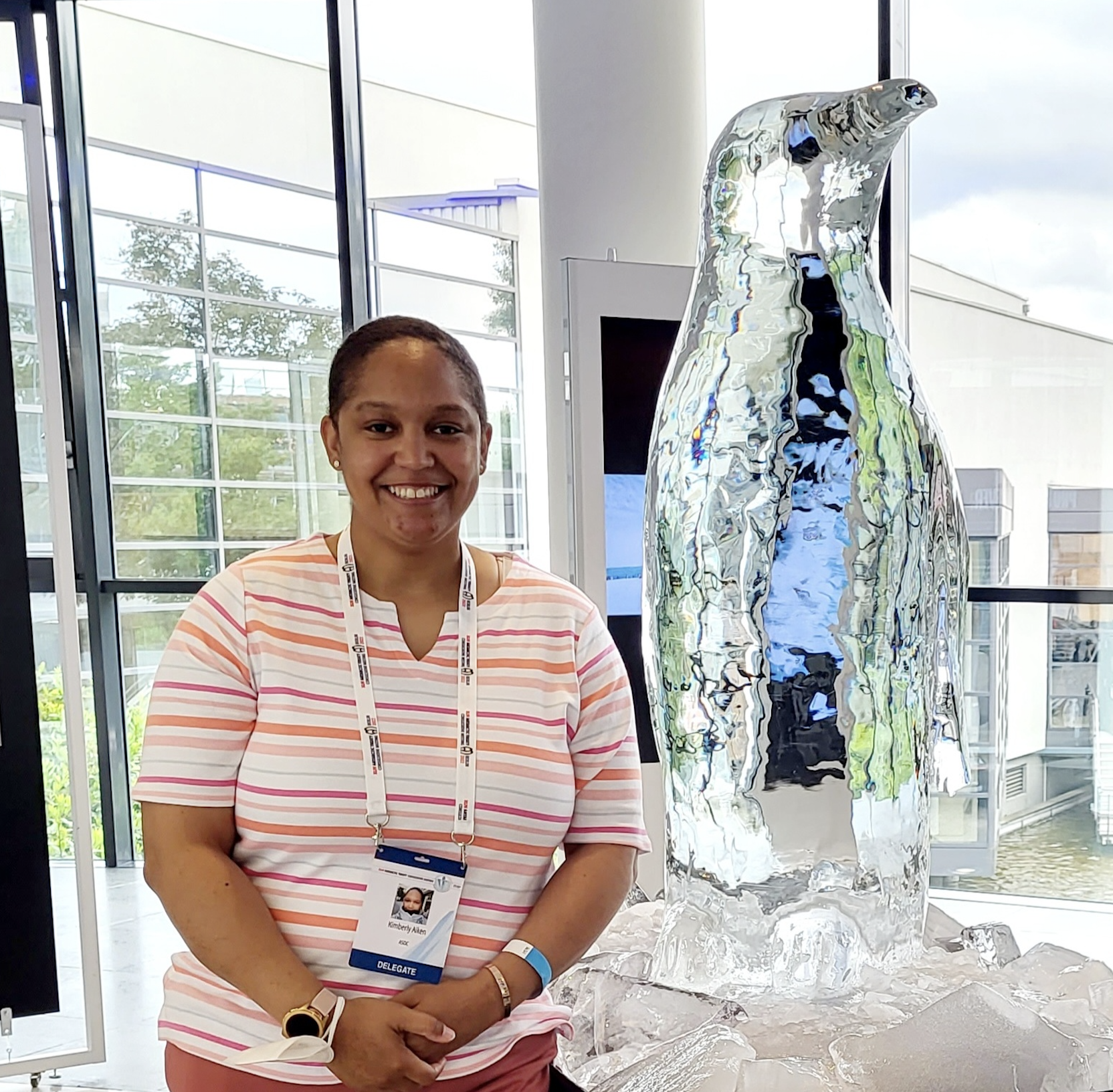 Kimberly Aiken, wearing an orange, white and pink striped shirt, pink trousers, and a lanyard stands beside an ice sculpture of a penguin