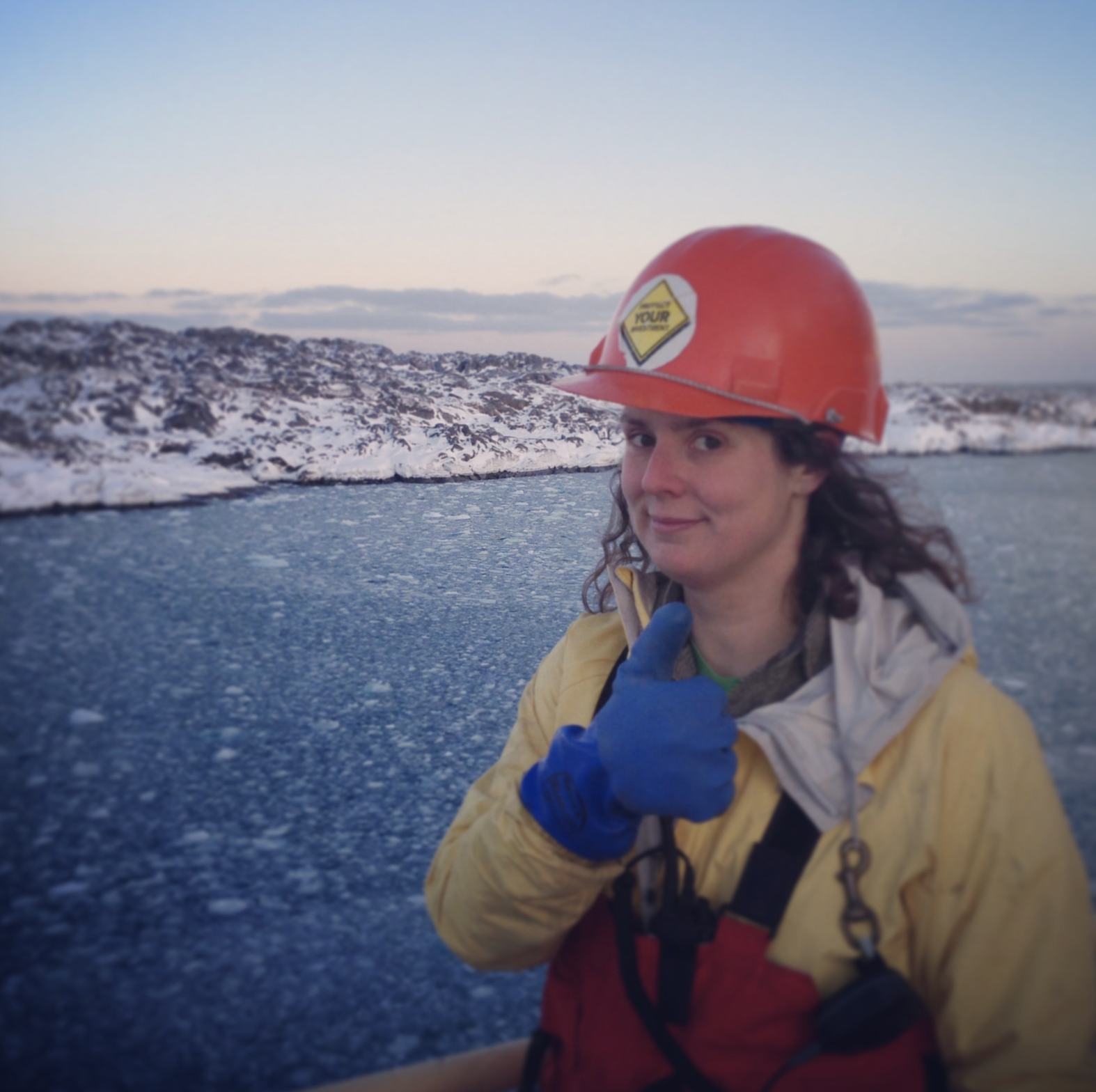 Jamee Johnson pictured wearing a hard hat on a vessel in Antarctica, looking at the camera and showing a thumbs up with a gloved hand