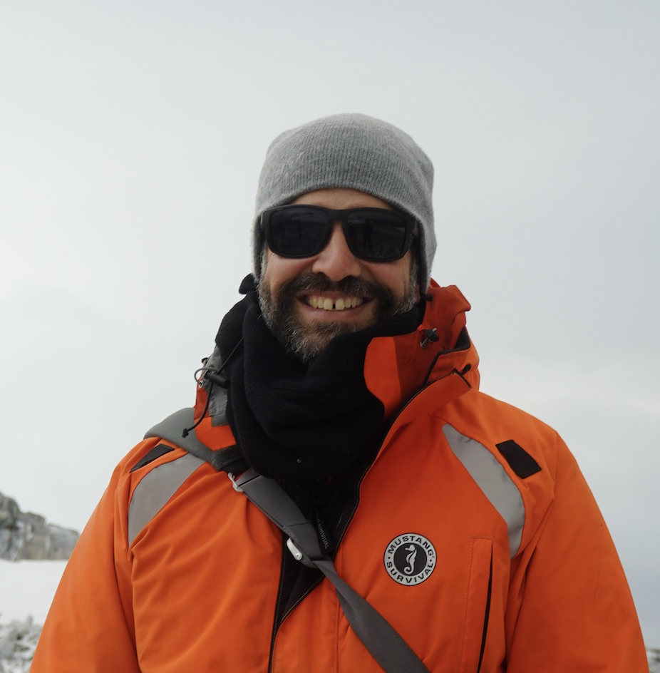 Carlos grins at the camera, bundled up in bright orange cold weather clothing in front of an icy landscape 