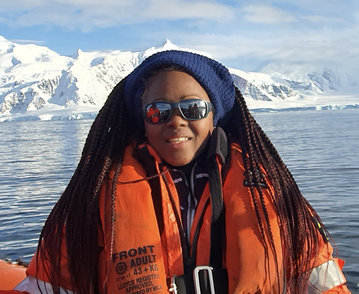 Nicole Logan-Park wears a bright orange life vest while on a boat, with a scene in the background of an icy fjord with mountains in the background.