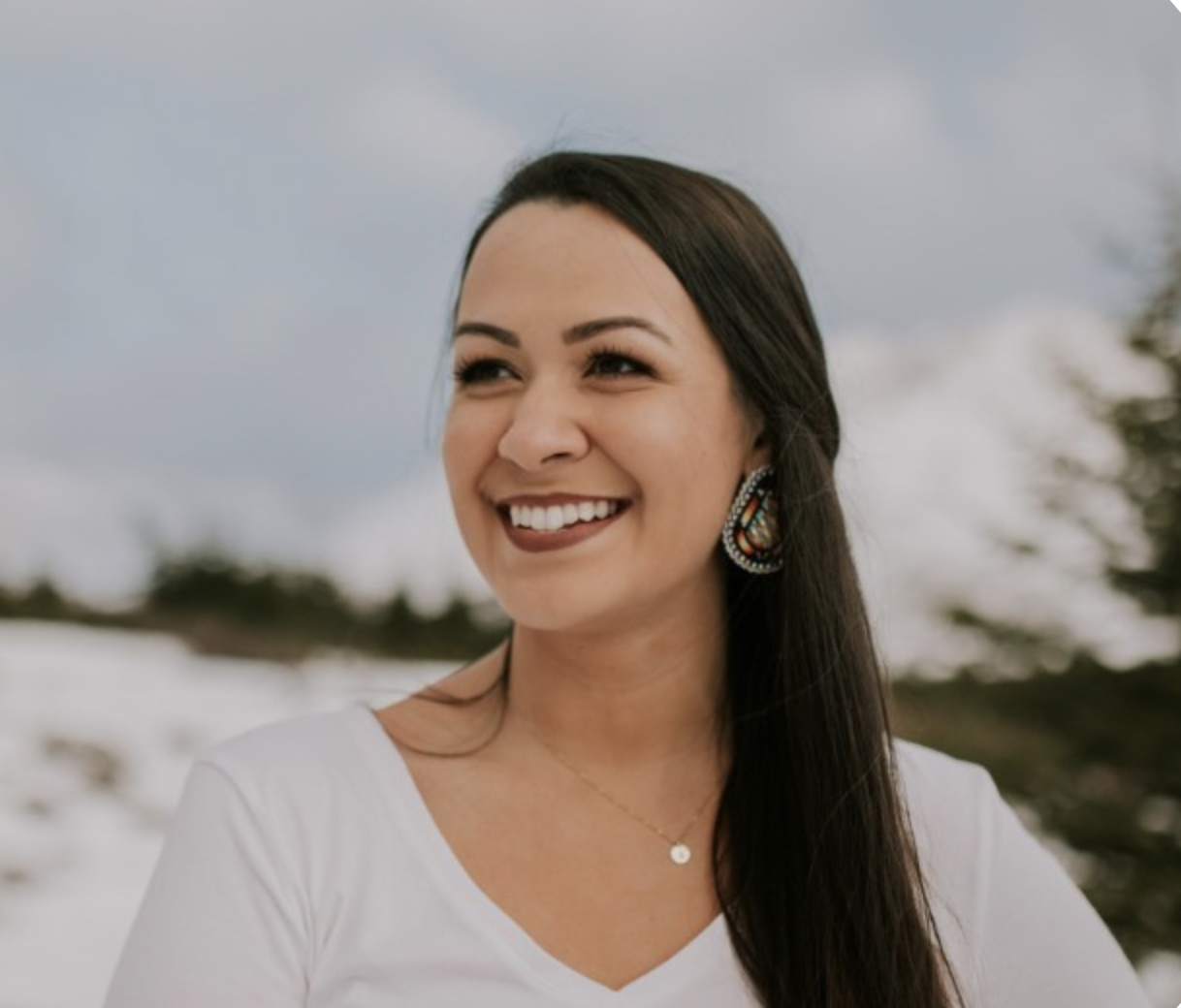 Karli Tyance Hassell (woman with brown hair, tan skin, wearing lipstick and earrings) gazes into middle distance - smiling. Karli is wearing a white top and has a mountain scene behind her.  