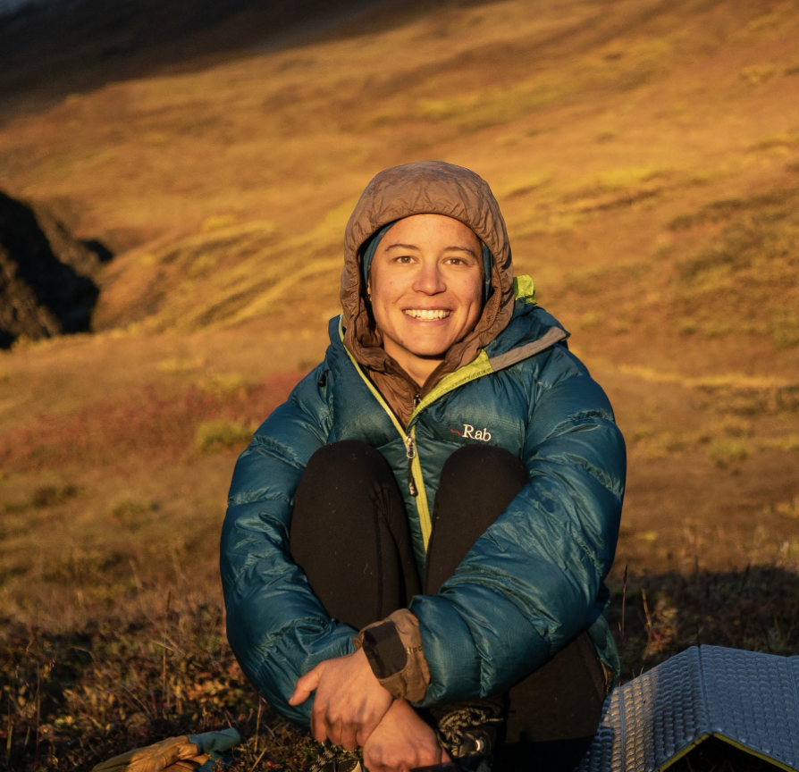 Kirsten (woman wearing two puffy jackets, black trousers with tan skin and brown hair) sits on the ground on golden colored grass and an accordion sleeping pad, smilling at the camera. 