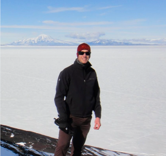Michael stands wearing a red hat and long sleeved jacket with an ice sheet in the background