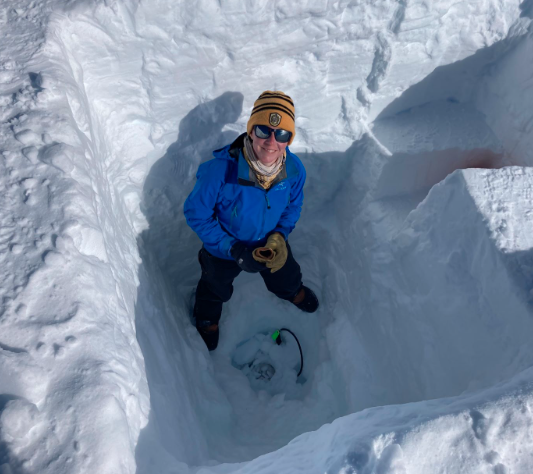 Marianne wears a yellow hat, blue jacket and stares up at the camera from within a snow pit