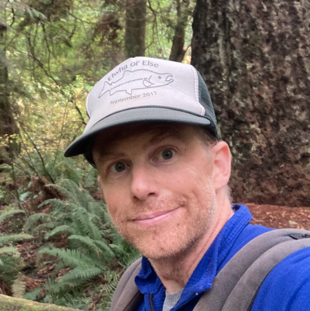Max wears a ball cap that says 'Elwha or Else | September 2017 and smiles at the camera from within a forest