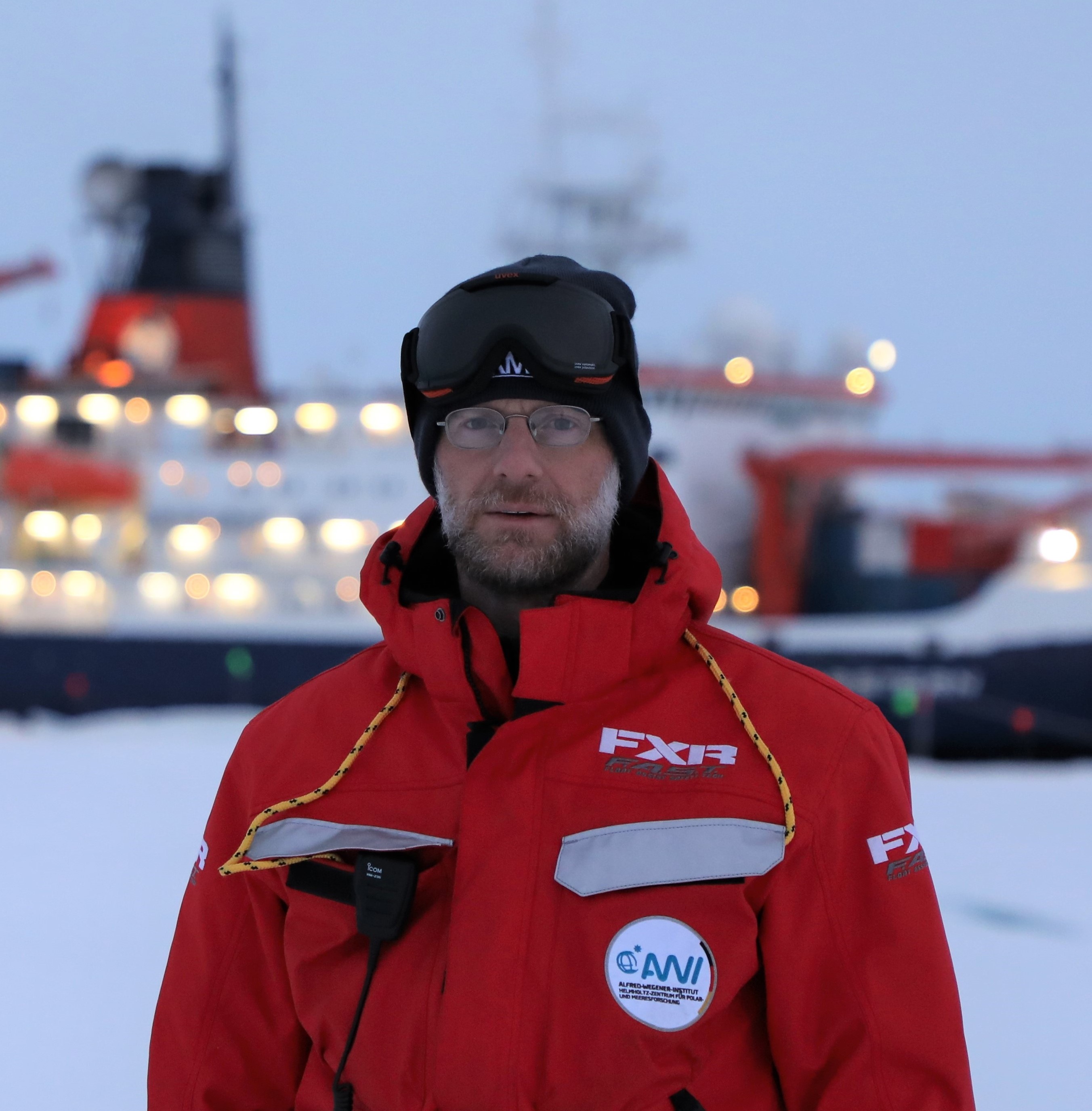 Matthew Shupe is all bundled up in winter weather gear and gazes at the camera on sea ice in front of an icebreaker ship in the Arctic