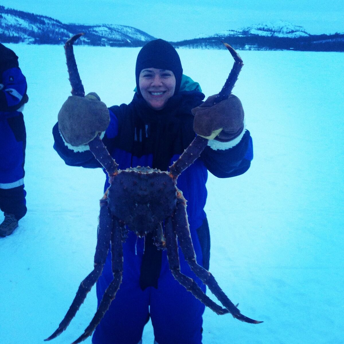Colleen holds a King Crab up to the camera in front of a snowy scene