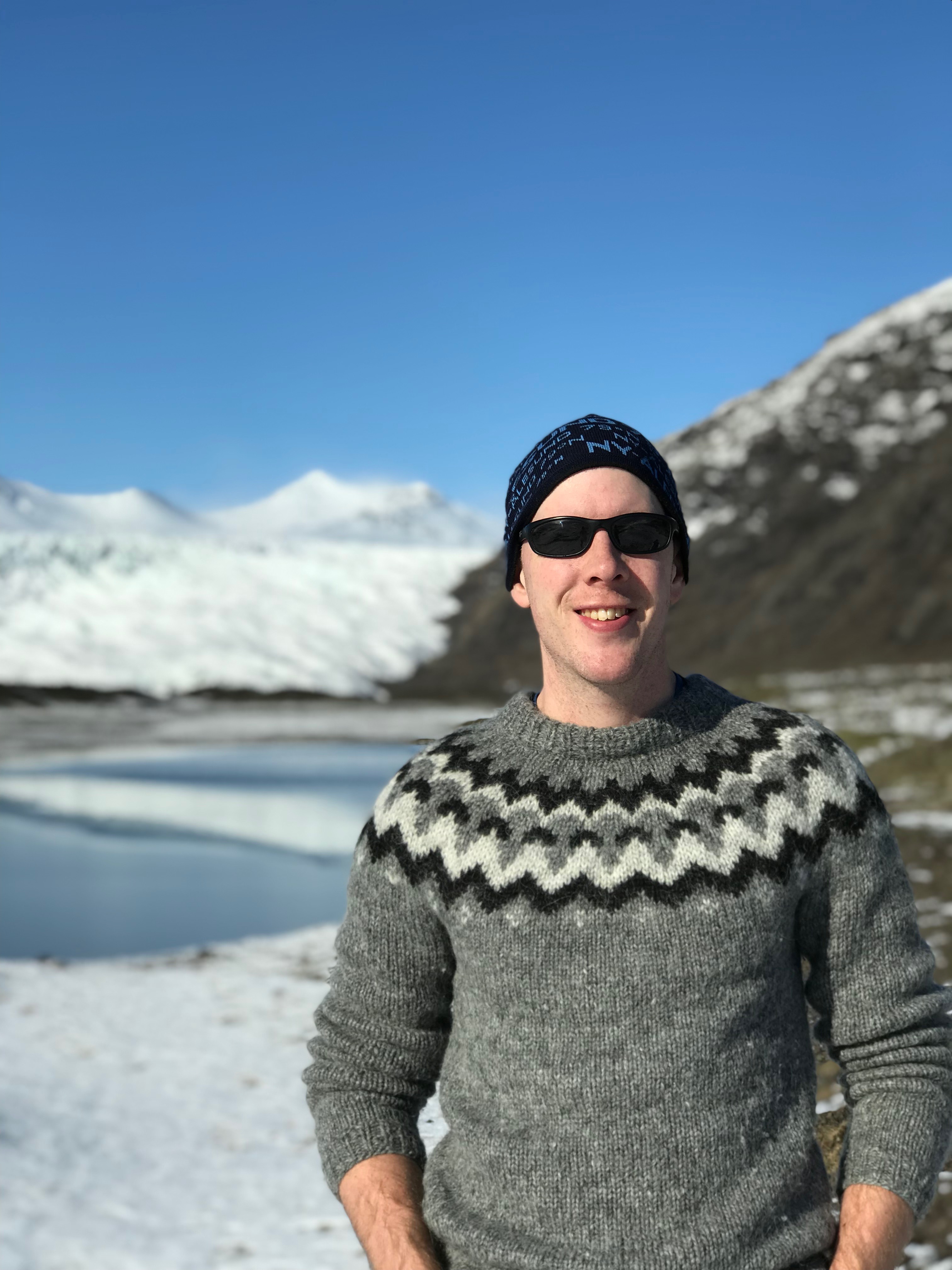 Allen stands in front of a mountainous and glaciated scene, wearing a classic Icelandic-patterned wool sweater, sunglasses and a hat.