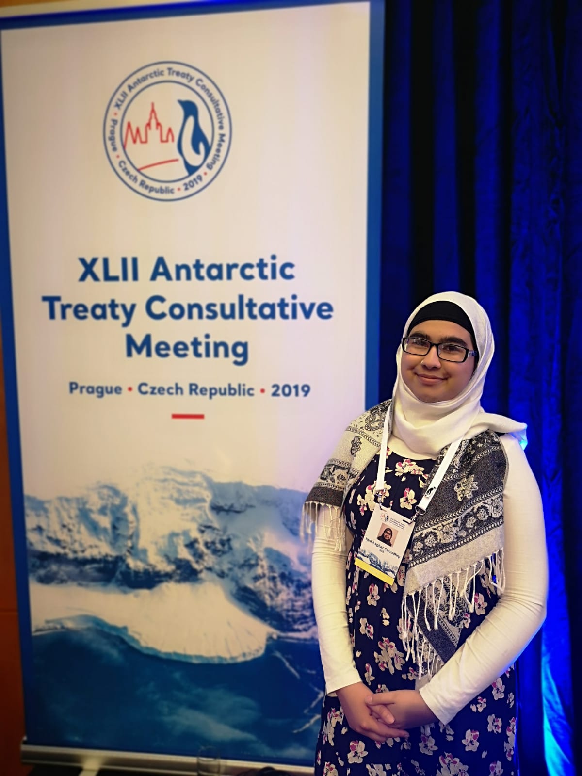 Dr. Iqra Choudhry stands beside a banner that reads 'XLII Antarctic Treaty Consultative Meeting, Prague, Czech Republic, 2019'.