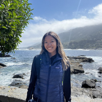 Rose Leeger outside on a beach. Rose is smiling at the camera with a rocky shore behind her. There is a tree on the left and mountain backdrop with clouds to the right. 