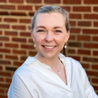 Marion McKenzie (a woman with dark blonde hair and light skin wearing earrings and a white blouse) smiles at the camera in front of a red brick wall