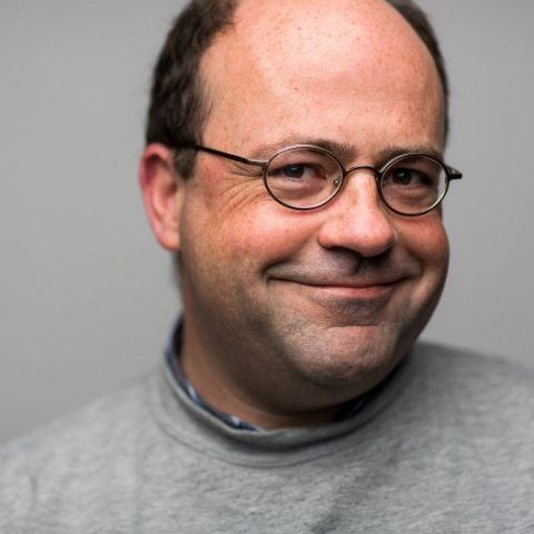 A man with black hair and stubble wearing glasses smiles. He is wearing a grey shirt and standing in front of a grey background. 
