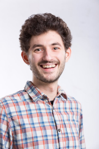 A man with short brown curly hair smiles in front of a white background. 
