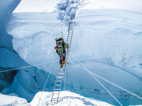 A woman traverses over a large gap in the ice. She is wearing a large backpack, orange snowsuit, and is walking on a ladder. 