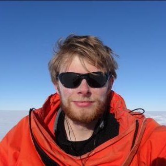 A blonde man wearing an orange jacket and sunglasses smiles at the camera. 