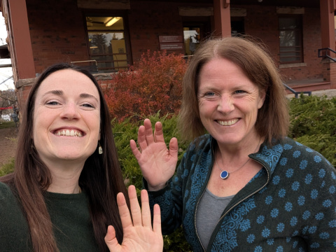 Mariama Dryák-Vallies, Director of PSECCO (woman with fair skin, dark brown hair and wearing a green sweater) smiles at the camera while waving next to Anne Gold, PI for PSECCO (woman with fair skin, brown hair and wearing a patterned sweater), who is also smiling at the camera and waving