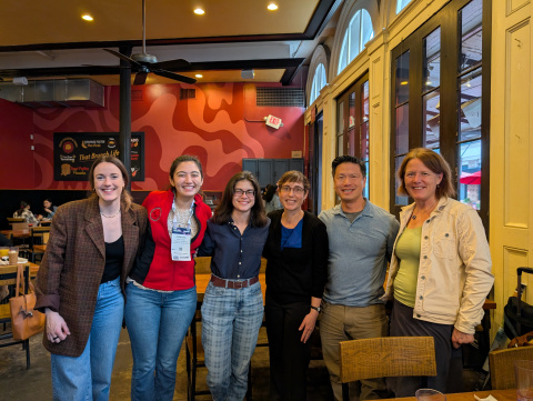 Photo of the PSECCO Advisory Board; pictured left to right is Mariama Dryák-Vallies (PSECCO Director), Asmara Lehrmann, Madison Payne (PSECCO Events Coordinator), Twila Moon, Gifford Wong, and Anne Gold. They stand inside a café with brightly painted walls behind them.