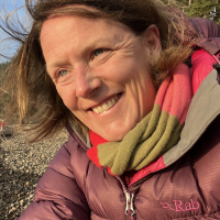 Person in a burgundy coat and a red and green scarf wrapped around their neck. Trees and gravel in the background.