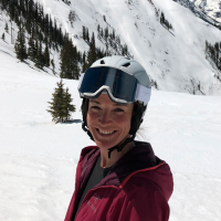 Person in a red jacket and white helmet with black goggles. A snow covered mountain and trees are in the background.