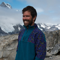 Person in a blue jacket with red, yellow and blue patterned sleeves standing in front of mountains covered in fog and snow.