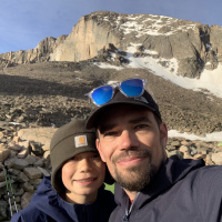 Person in front: Adult in a brown hat with blue sunglasses and a black jacket. Person in the back: child in a blue jacket and a brown hat. A hill and rocks are in the background.