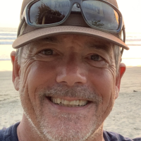 Person in a blue t-shirt, brown hat and black sunglasses. A beach in the background.