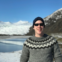 Person in a black hat and black sunglasses wearing a grey sweater with black and white zig zag stripes. A lake in the background, as well as snow on the ground and snow-covered mountains.