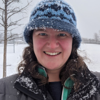 Person in a green jacket, black coat and blue hat. A tree with no leaves in the background, as well as snow on the ground.