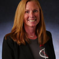 Person in a grey shirt with a red and white logo and a black blazer. Background is a solid white at the bottom and fades into grey and black at the top of the image.