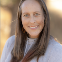 Person with long brown hair in a grey hoodie. Trees blurred into the background.