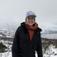 Person in a red shirt, black coat and blue hat with a red scarf around their neck. A background of trees, snow, and mountains covered in fog.