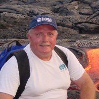 A man with a white shirt, blue hat, and blue backpack smiles in front of a flowing volcano.