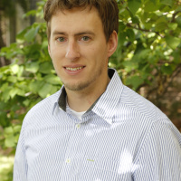 A man in a seersucker button-down shirt smiles in front of green foliage. 