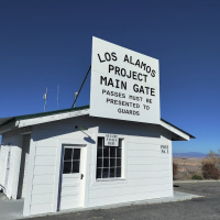 A large white building stands with a sign stating "Los Alamos Project Main Gate - passes must be presented to guards" 