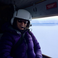 Jessie Crain sits in a helicopter with a helmet, sunglasses, puffer jacket, and a big smile