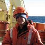 Michelle poses outside on a research vessel ship, wearing a hard hat and a big orange jacket