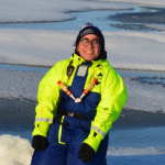 Alex (a woman with dark brown hair and tanned skin, wearing a bobble hat, bright yellow jacket and blue overalls) kneels on seal ice doing science. 