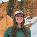 Mikaela Williams stands in front of bright orange rock with snow on it, wearing a green long-sleeved top and a white hat, grinning at the camera