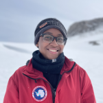 Maya Thomas stands in Antarctica wearing a warm hat, glasses, a buff and a red 'USAP NSF' jacket