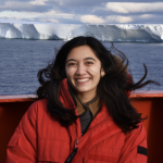 Asmara Lehrmann stands on a boat in front of a big ice shelf in Antarctica with ocean in between. Asmara wears a big red jacket and smiles at the camera. 