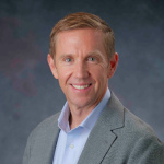 Steve Lommele (white man with light brown hair and grey eyes) smiles at the camera. Steve is wearing a collared shirt and suit jacket. 